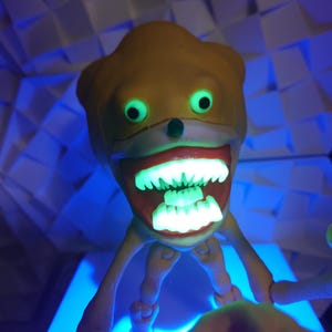 May include: A yellow and white toy with a large toothy grin and glowing green eyes. The toy has long, thin arms and legs.