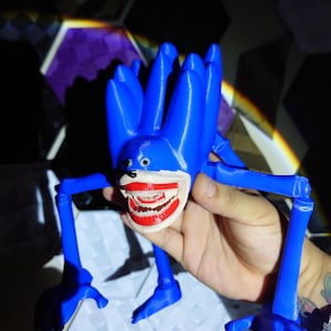 May include: A blue, 3D-printed figure with a large, open mouth revealing teeth. The figure has multiple appendages and a cartoonish design. The figure is held in a hand.
