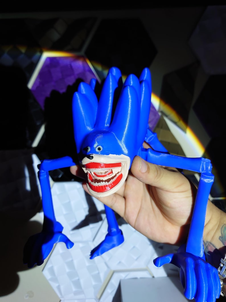 May include: A blue, 3D-printed figure with a large, open mouth revealing teeth. The figure has multiple appendages and a cartoonish design. The figure is held in a hand.