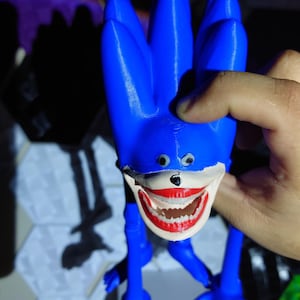 May include: A blue, 3D-printed figurine of a character with a large, unsettling grin. The figure has long, spiky hair, small eyes, and elongated limbs. The mouth is painted red with white teeth. The figurine is held by a hand.