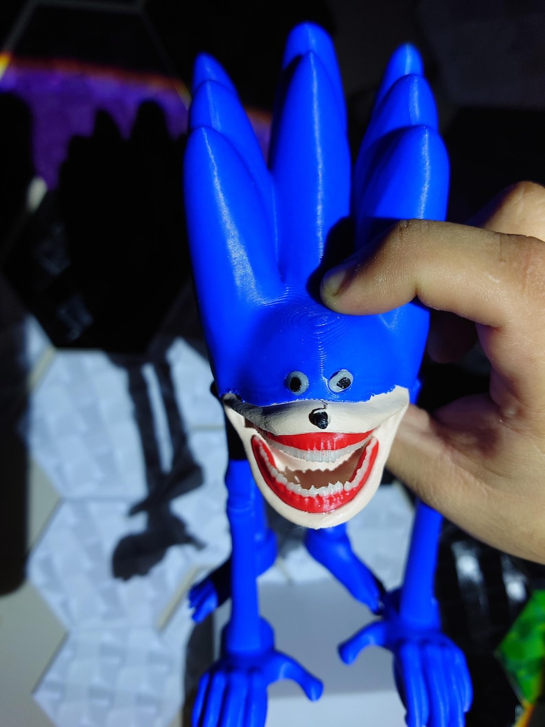 May include: A blue, 3D-printed figurine of a character with a large, unsettling grin. The figure has long, spiky hair, small eyes, and elongated limbs. The mouth is painted red with white teeth. The figurine is held by a hand.