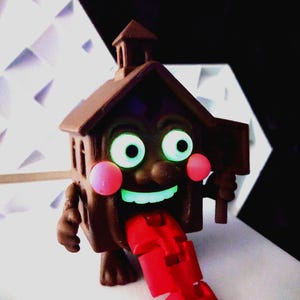 May include: A brown, house-shaped toy with glowing green eyes and a smiling mouth. The toy has pink cheeks and a red, segmented tongue extending out. It is holding a small sign.