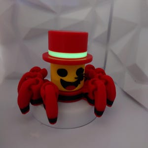 Op de afbeelding: Een grillig rood en geel spinnenbeeldje met een hoge hoed. De spin heeft acht rode poten met zwarte uiteinden, een lachend gezicht en een zwarte streep rond zijn lichaam. De hoge hoed is rood met een groene band.