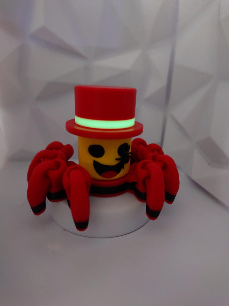 Op de afbeelding: Een grillig rood en geel spinnenbeeldje met een hoge hoed. De spin heeft acht rode poten met zwarte uiteinden, een lachend gezicht en een zwarte streep rond zijn lichaam. De hoge hoed is rood met een groene band.