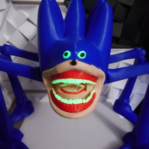 May include: A blue, spiky-haired creature with glowing green eyes and teeth. The creature has multiple blue legs and a large, open mouth with red lips. The teeth glow green, creating a striking contrast against the blue and tan colors.