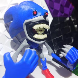 Puede incluir: Una figura azul y gris de un personaje con una gran sonrisa dentuda y ojos rojos. La figura tiene manos y pies azules y está posada sobre un fondo geométrico. La figura es una representación estilizada de un popular personaje de videojuego.