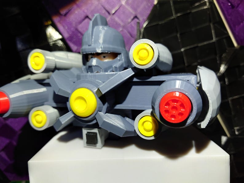 May include: A gray plastic toy robot with a helmet and multiple yellow and red cannons. The robot is posed with its cannons facing forward.