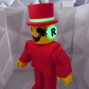 Op de afbeelding: Een rode figuur met hoge hoed en pak, gele gezicht en handen. De figuur heeft een groen oplichtende cirkel op zijn gezicht met de letter "R" en een zwarte spin. Het staat op een witte basis.