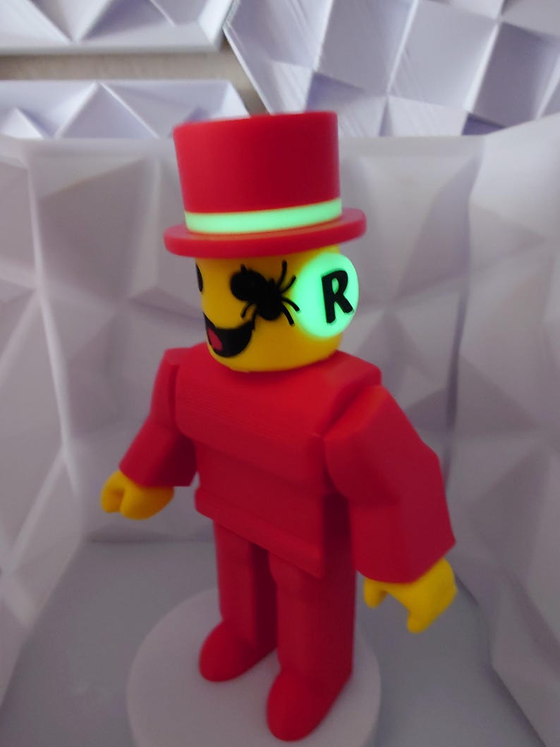 Op de afbeelding: Een rode figuur met hoge hoed en pak, gele gezicht en handen. De figuur heeft een groen oplichtende cirkel op zijn gezicht met de letter "R" en een zwarte spin. Het staat op een witte basis.