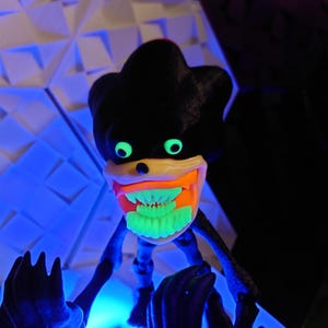 May include: A black and green toy figure with a large, toothy grin. The figure has glowing green eyes and teeth, and is posed with its arms outstretched.