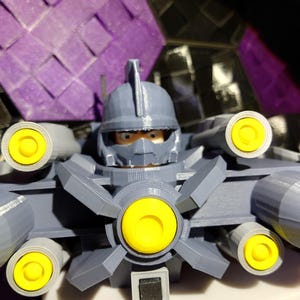 May include: A gray 3D printed toy robot with yellow details and a black button on the front. The robot has a helmet with a face and multiple cannons on the sides.