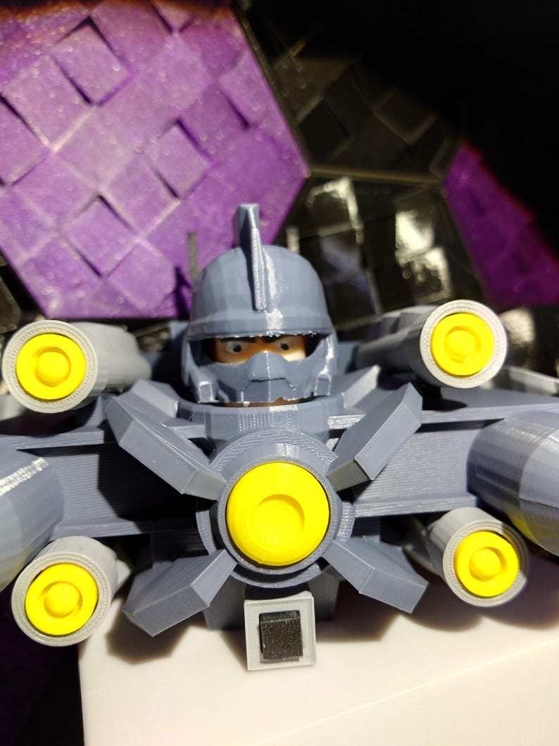 May include: A gray 3D printed toy robot with yellow details and a black button on the front. The robot has a helmet with a face and multiple cannons on the sides.