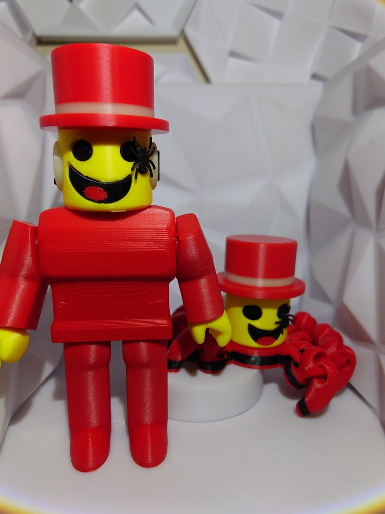 Op de afbeelding: Een rood, geleed figuur met een gele kop, zwarte ogen en een lachende mond, met een rode hoge hoed. Een kleine zwarte spin is aan het gezicht van het figuur bevestigd. Een ander vergelijkbaar figuur staat op de achtergrond.