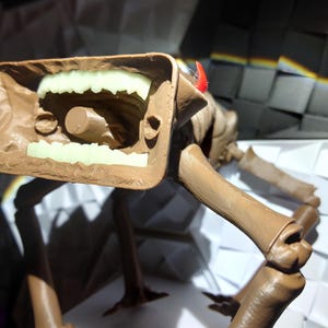 May include: A brown, articulated creature figurine with a large, open mouth revealing glowing green teeth. The figure has multiple legs and a small red horn. The design appears to be a stylized monster or alien.