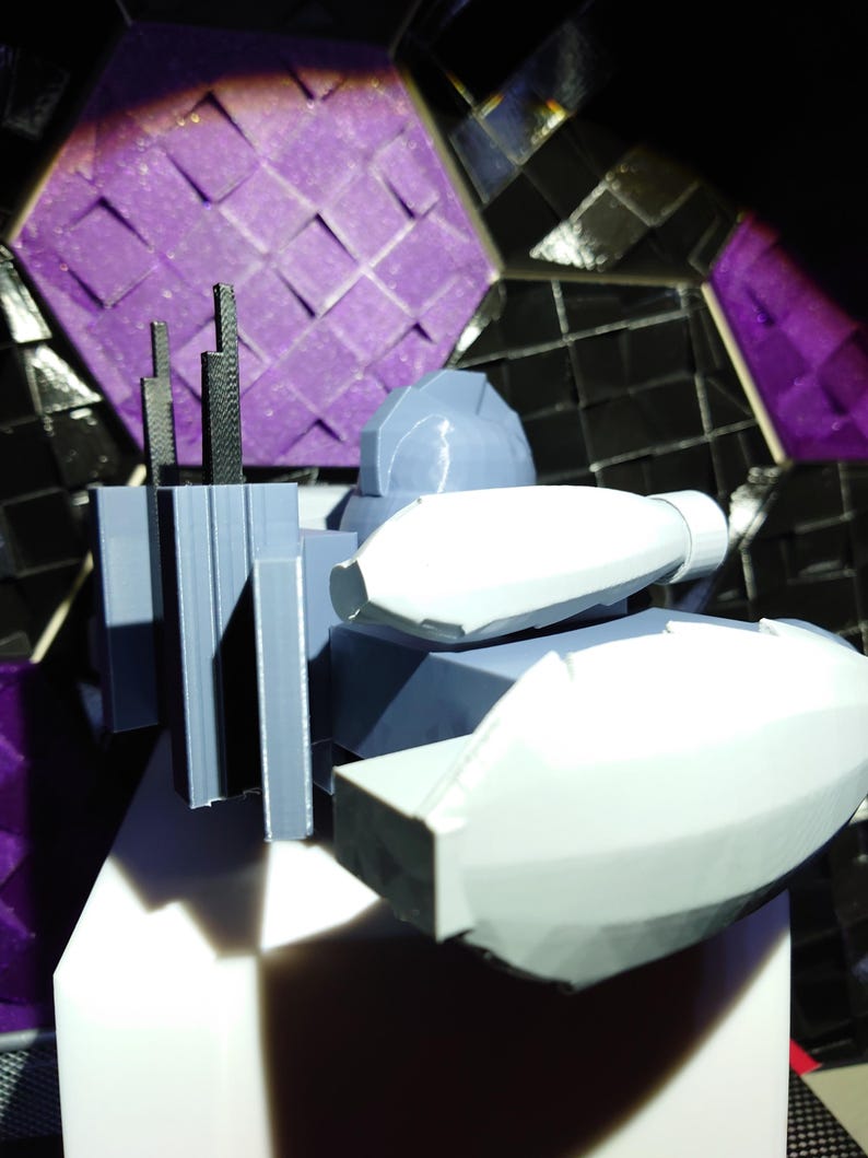 May include: A 3D printed model of a futuristic spaceship with a gray and white color scheme. The spaceship has a large, rounded front section and a smaller, rectangular rear section. There are several antennas on the top of the spaceship.