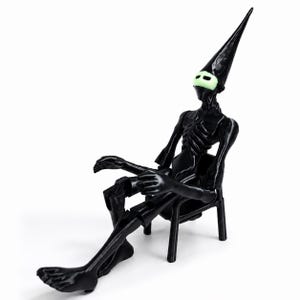 May include: A black articulated figurine with a tall, pointed headpiece and a pale, glowing mask featuring two horizontal eye slits. The figure has a skeletal torso and is seated on a small, matching black chair with four legs and a backrest.