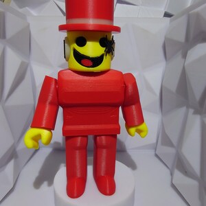 Op de afbeelding: Een 3D-geprint figuur van een persoon met een gele kop, rode lichaam en hoge hoed. Het figuur heeft een lachend gezicht en een kleine zwarte spin aan de zijkant van zijn hoofd. Het figuur staat op een witte, cirkelvormige basis.