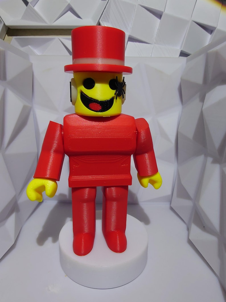 Op de afbeelding: Een 3D-geprint figuur van een persoon met een gele kop, rode lichaam en hoge hoed. Het figuur heeft een lachend gezicht en een kleine zwarte spin aan de zijkant van zijn hoofd. Het figuur staat op een witte, cirkelvormige basis.
