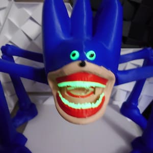 May include: A blue, cartoonish creature with glowing green eyes and a wide, red-lipped mouth. The creature has multiple blue appendages extending from its body, giving it a spider-like appearance. The mouth and teeth also glow green.