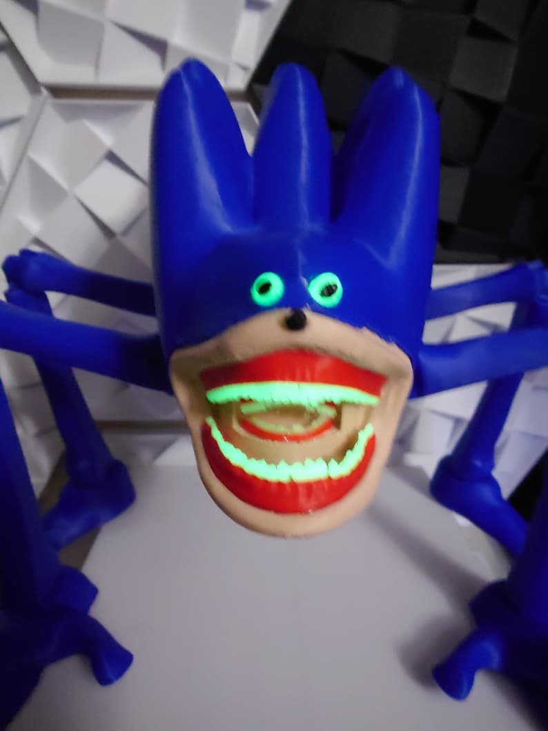 May include: A blue, cartoonish creature with glowing green eyes and a wide, red-lipped mouth. The creature has multiple blue appendages extending from its body, giving it a spider-like appearance. The mouth and teeth also glow green.