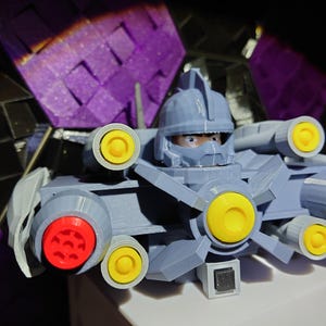 May include: A gray 3D printed robot toy with yellow and red details. The robot has a head with a visor and a large gun on the side.