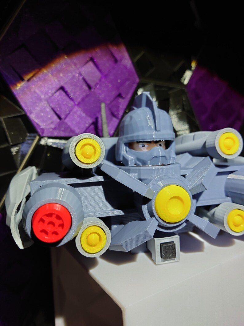 May include: A gray 3D printed robot toy with yellow and red details. The robot has a head with a visor and a large gun on the side.