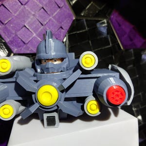 May include: A gray 3D printed robot toy with yellow and red details. The robot has a helmet, a large gun, and a spinning fan.