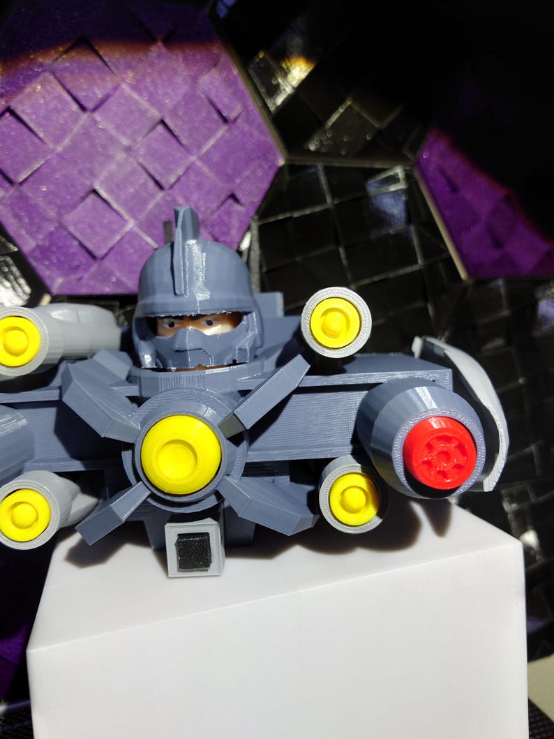 May include: A gray 3D printed robot toy with yellow and red details. The robot has a helmet, a large gun, and a spinning fan.