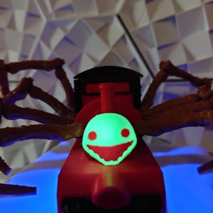 Könnte beinhalten: Ein roter Spielzeugzug mit einem grün leuchtenden Gesicht und einem grinsenden Gebiss. Der Zug hat braune spinnenartige Beine.