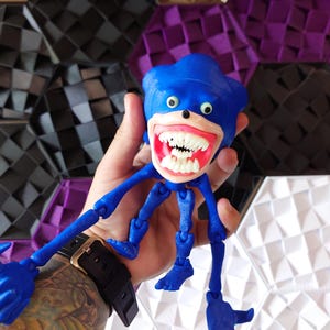 May include: A blue articulated toy figure of Sonic the Hedgehog with a wide, toothy grin and large, white eyes. The figure has multiple joints and is posed with its arms and legs outstretched.
