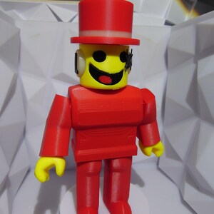 Op de afbeelding: Een rood en geel speelfiguur met een hoge hoed. Het figuur heeft een gele kop met een lachend gezicht en zwarte ogen. Het lichaam, de armen en benen zijn rood. Het figuur staat op een witte basis.