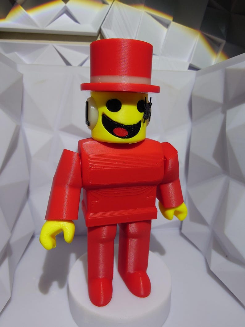 Op de afbeelding: Een rood en geel speelfiguur met een hoge hoed. Het figuur heeft een gele kop met een lachend gezicht en zwarte ogen. Het lichaam, de armen en benen zijn rood. Het figuur staat op een witte basis.