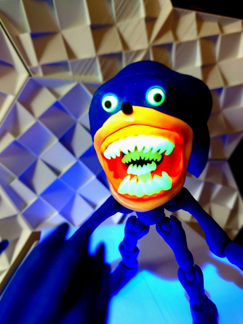 May include: A blue and orange articulated toy figure of a cartoon character with a wide open mouth and large teeth. The figure has a blue body, orange mouth, and green eyes.