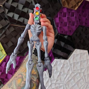 May include: A gray articulated skeleton figure with a traffic light head. The traffic light has red, yellow, and green lights. The figure is held in a person's hand.