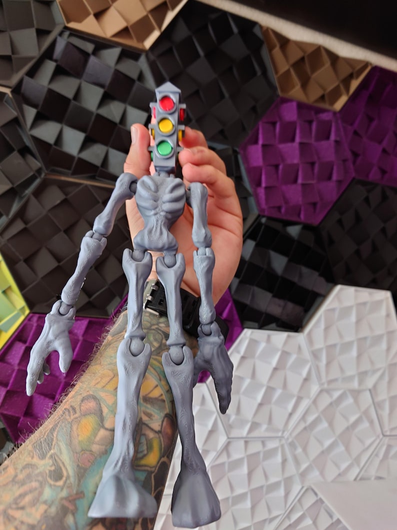 May include: A gray articulated skeleton figure with a traffic light head. The traffic light has red, yellow, and green lights. The figure is held in a person's hand.