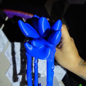 May include: A vibrant blue, 3D-printed sculpture of a stylized flower. The sculpture features a detailed floral design with elongated petals and stem-like supports. The piece is held in a hand, showcasing its size and form.