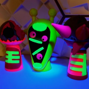 Op de afbeelding: Drie neon-groene, roze en zwarte alien-achtige figuren met gloeiende ogen. De figuren zijn gemaakt van plastic en hebben een cartoonachtig uiterlijk.
