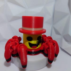 Op de afbeelding: Een 3D-geprint speelgoedspin met een gele kop, zwarte ogen en een lachende mond. Het draagt een rode hoge hoed en heeft rode poten met zwarte uiteinden. De spin staat op een witte basis.