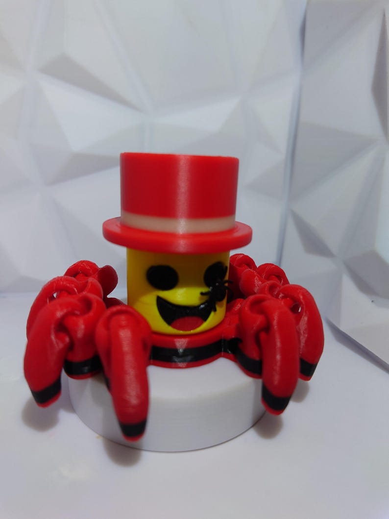 Op de afbeelding: Een 3D-geprint speelgoedspin met een gele kop, zwarte ogen en een lachende mond. Het draagt een rode hoge hoed en heeft rode poten met zwarte uiteinden. De spin staat op een witte basis.