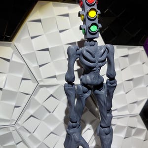May include: A gray articulated action figure with a traffic light head. The traffic light has red, yellow, and green lights.