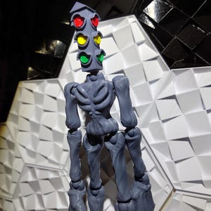 May include: A gray articulated figure with a traffic light for a head. The traffic light has red, yellow, and green lights. The figure is standing on a white and black patterned surface.