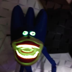 May include: A blue, cartoon-like figure with glowing green eyes and a wide, grinning mouth. The mouth and teeth glow with a bright green light. The figure has a three-pronged head and thin, blue limbs.
