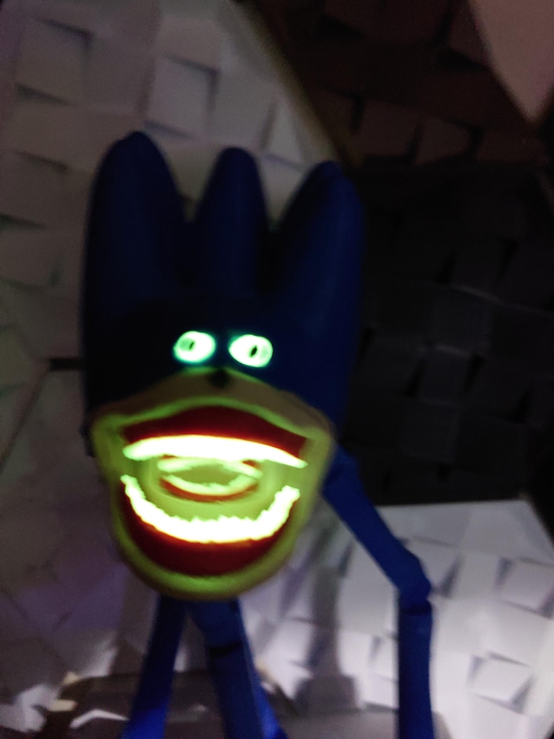 May include: A blue, cartoon-like figure with glowing green eyes and a wide, grinning mouth. The mouth and teeth glow with a bright green light. The figure has a three-pronged head and thin, blue limbs.