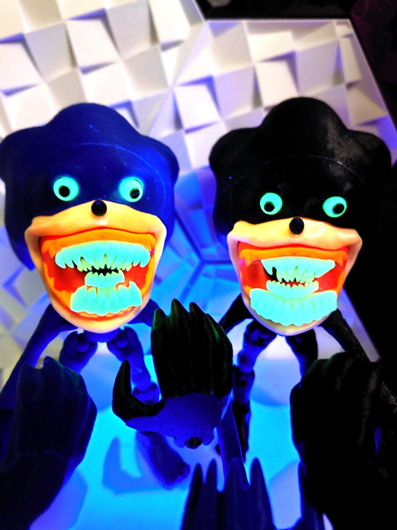 May include: Two black and blue figures with glowing green eyes and teeth. The figures are posed with their arms outstretched and their hands in fists. The figures are set against a white background.