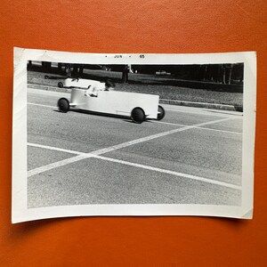 May include: A black and white photograph of a homemade soapbox derby car. The car is white and has a black steering wheel. The car is on a paved road with white lines. The photo is dated 