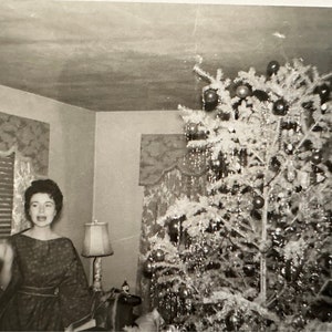 May include: A woman in a patterned dress stands in front of a decorated Christmas tree. The tree is decorated with ornaments and tinsel.