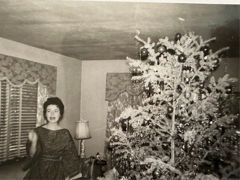 May include: A woman in a patterned dress stands in front of a decorated Christmas tree. The tree is decorated with ornaments and tinsel.