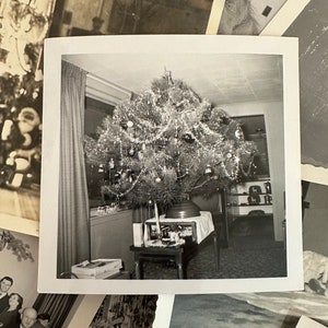 May include: A black and white photograph of a Christmas tree decorated with ornaments and tinsel. The tree is in a living room with a table and chairs in the background.
