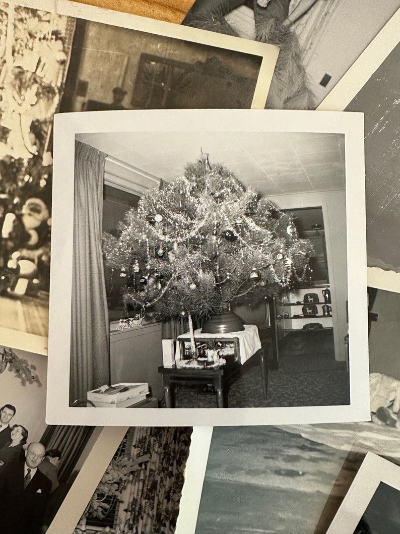 May include: A black and white photograph of a Christmas tree decorated with ornaments and tinsel. The tree is in a living room with a table and chairs in the background.