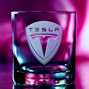 May include: A clear glass tumbler with the Tesla logo etched into the side. The logo is a white shield with the word "TESLA" above a stylized "T".